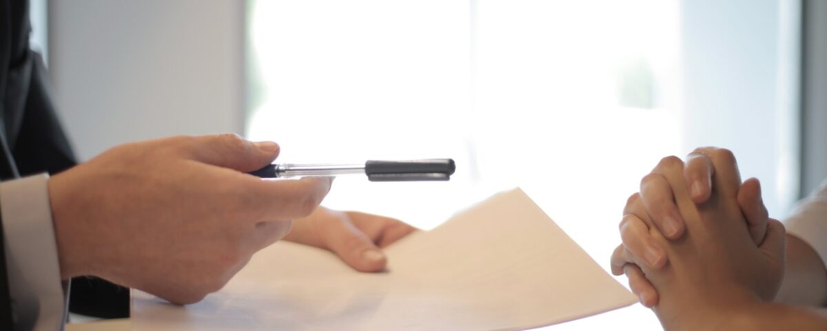 on left a set of hands holding an ink pen and paper handing it to another set of folded hands on the right.