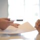 on left a set of hands holding an ink pen and paper handing it to another set of folded hands on the right.