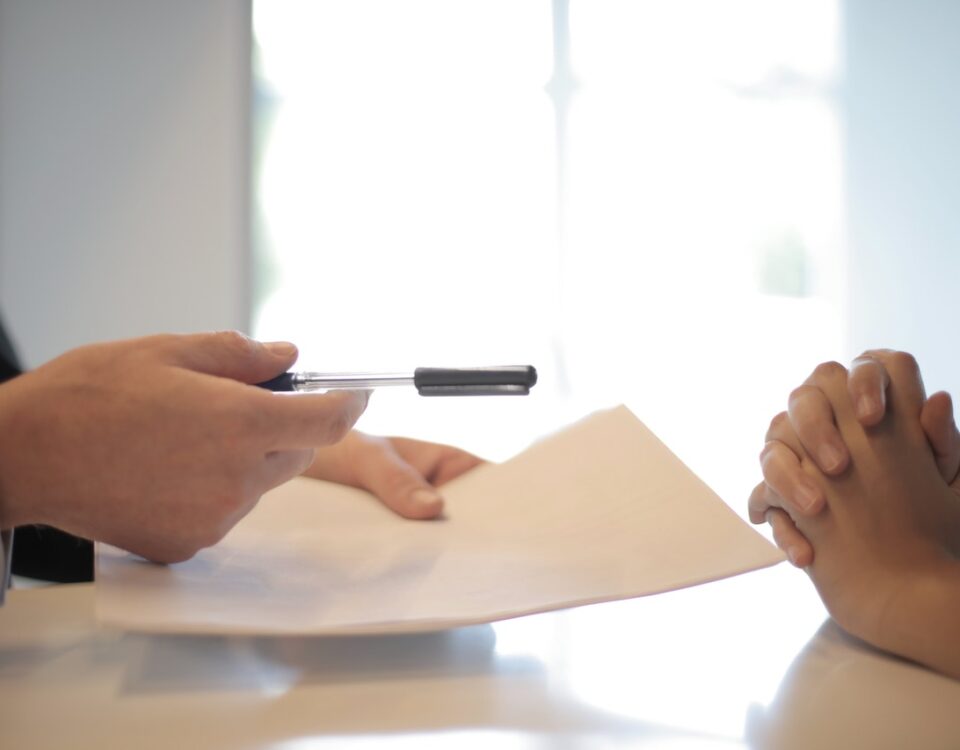 on left a set of hands holding an ink pen and paper handing it to another set of folded hands on the right.