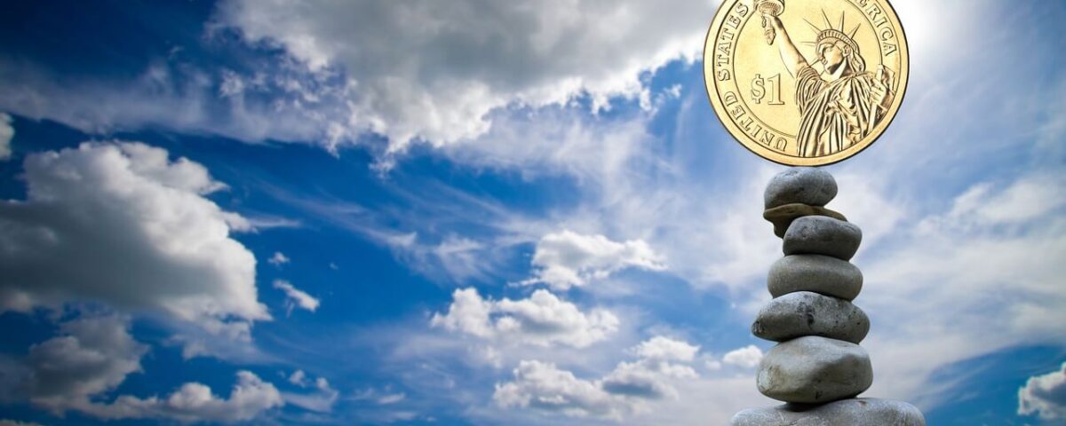 blue sky with fluffy white clouds. On the right is a stack of 7 river rocks stacked atop one another. At the top is a US gold $1 coin with the Statue of Liberty on it.