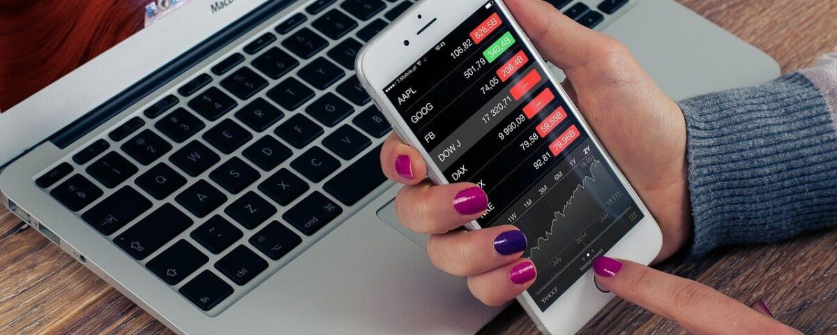 Mac Book Air on a desk. a woman's hand with purple nails holding a cell phone. on the phone appears to be stock market information