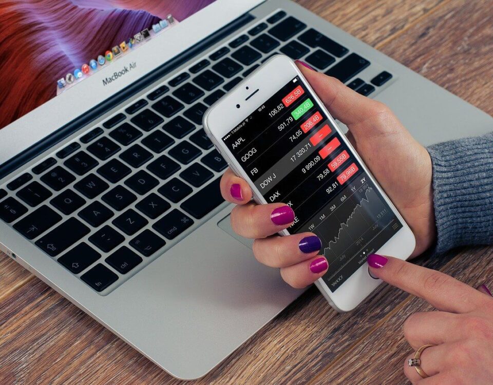 Mac Book Air on a desk. a woman's hand with purple nails holding a cell phone. on the phone appears to be stock market information