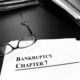 black and white image of file folders on a desk, a pair of glasses laying on a sheet of paper that reads "Bankruptcy Chapter 7"