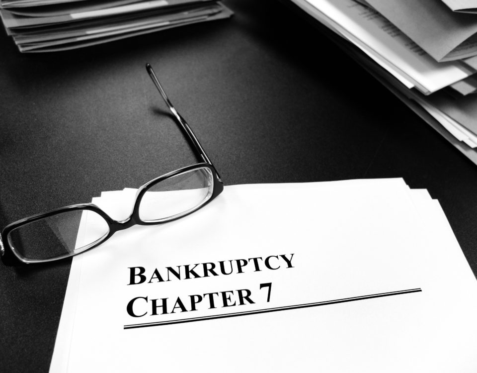 black and white image of file folders on a desk, a pair of glasses laying on a sheet of paper that reads "Bankruptcy Chapter 7"