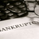 paper with "Bankruptcy" In bold black letters sticking out of a file folder, a calculator with a pair of black-rimmed glasses on top of it